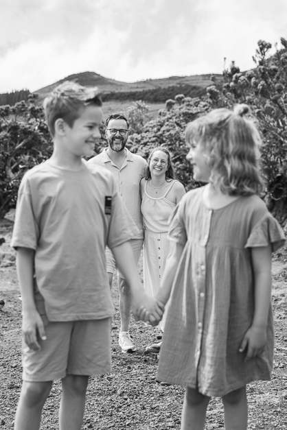 Black and white image of a family of four where the kids are in front of the camera holding hands, and their parents, behind of them, look smiling towards the kids